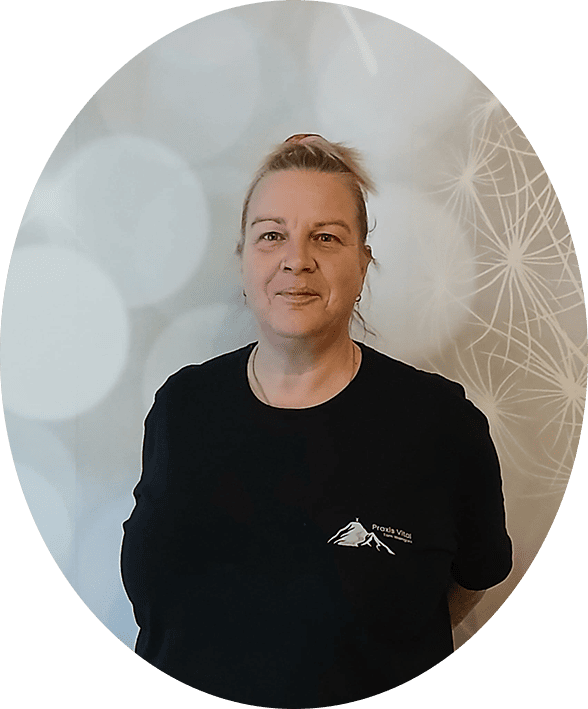 Person mit hochgebundenem Haar trägt schwarzes T-Shirt mit kleinem weißen Berglogo und Schriftzug 'Praxis am Berg' vor hellgrauer Wand mit weißem Pusteblumenmuster.